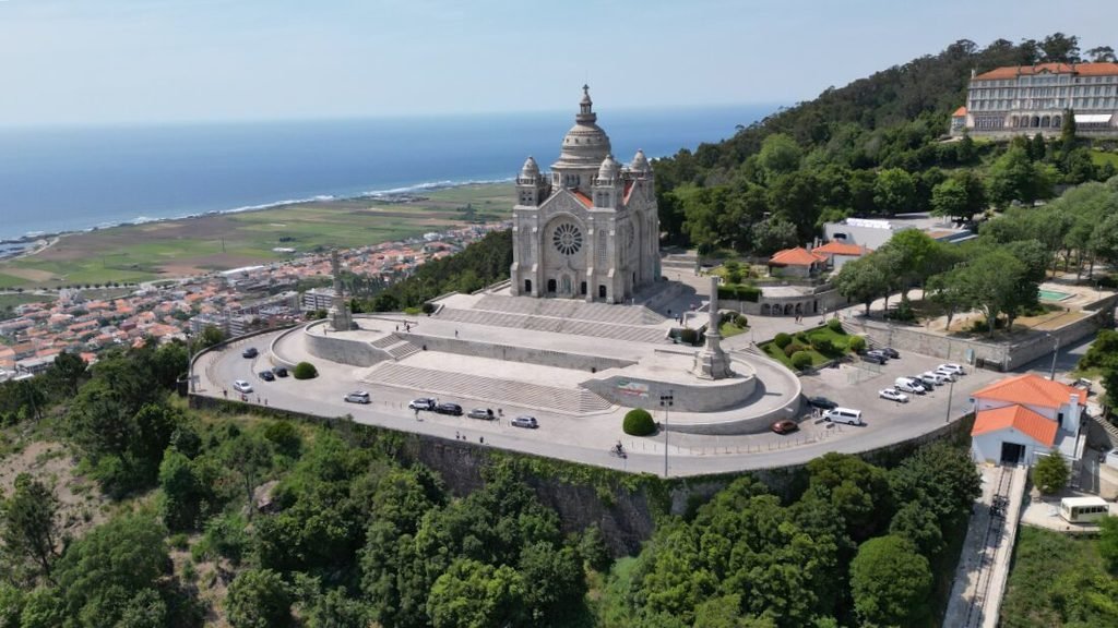 Best Towns in Northern Portugal: A Photographer’s Map A dramatic high-angle drone aerial photograph of the Sanctuary of Santa Luzia in Viana do Castelo, showcasing the grand Neo-Byzantine basilica on its massive stone terrace platform overlooking the green forested hills, coastal town, and blue Atlantic ocean horizon.