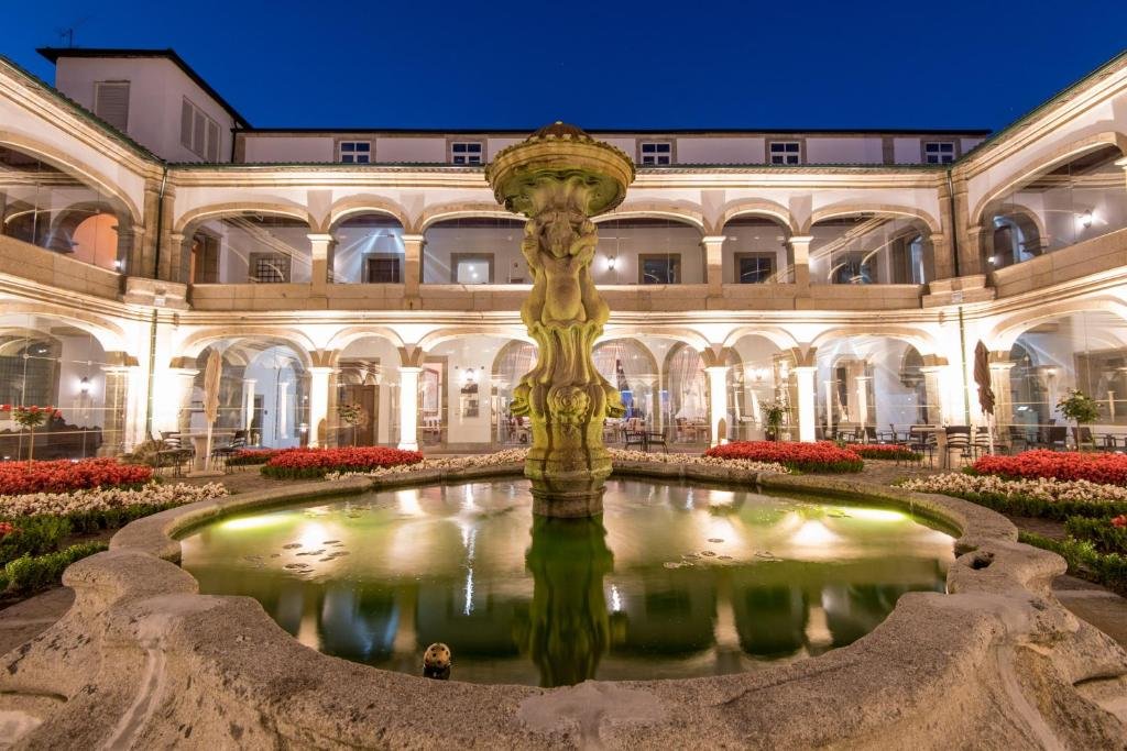 Best Towns in Northern Portugal: A Photographer’s Map An illuminated stone courtyard fountain at the Vila Galé Collection Braga hotel at night, featuring a multi-tiered baroque sculpture, surrounding flowerbeds, and a two-story arched building arcade with warm interior lighting.