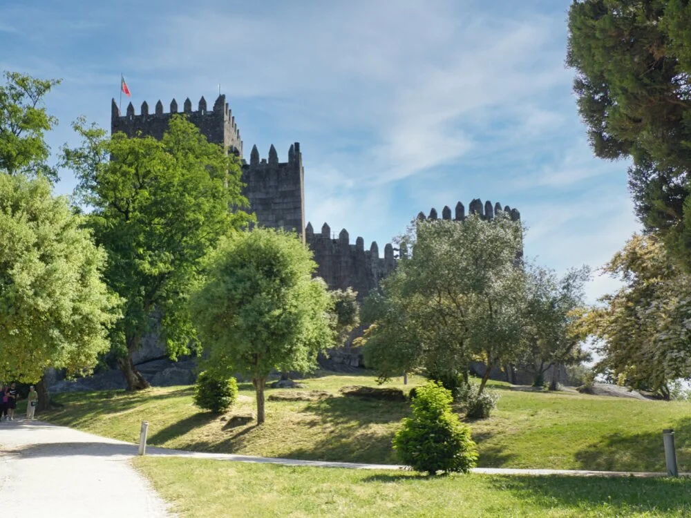 Guimarães Travel Guide: The Cradle of Stone and Soul A dirt walking path leading up to the historic stone towers of Guimarães Castle, framed by green trees and grass lawns under a partly cloudy sky.