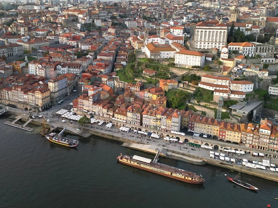 An aerial drone view of the Ribeira waterfront in Porto, Portugal. Traditional boats float along the Douro River next to historic red-roofed buildings.